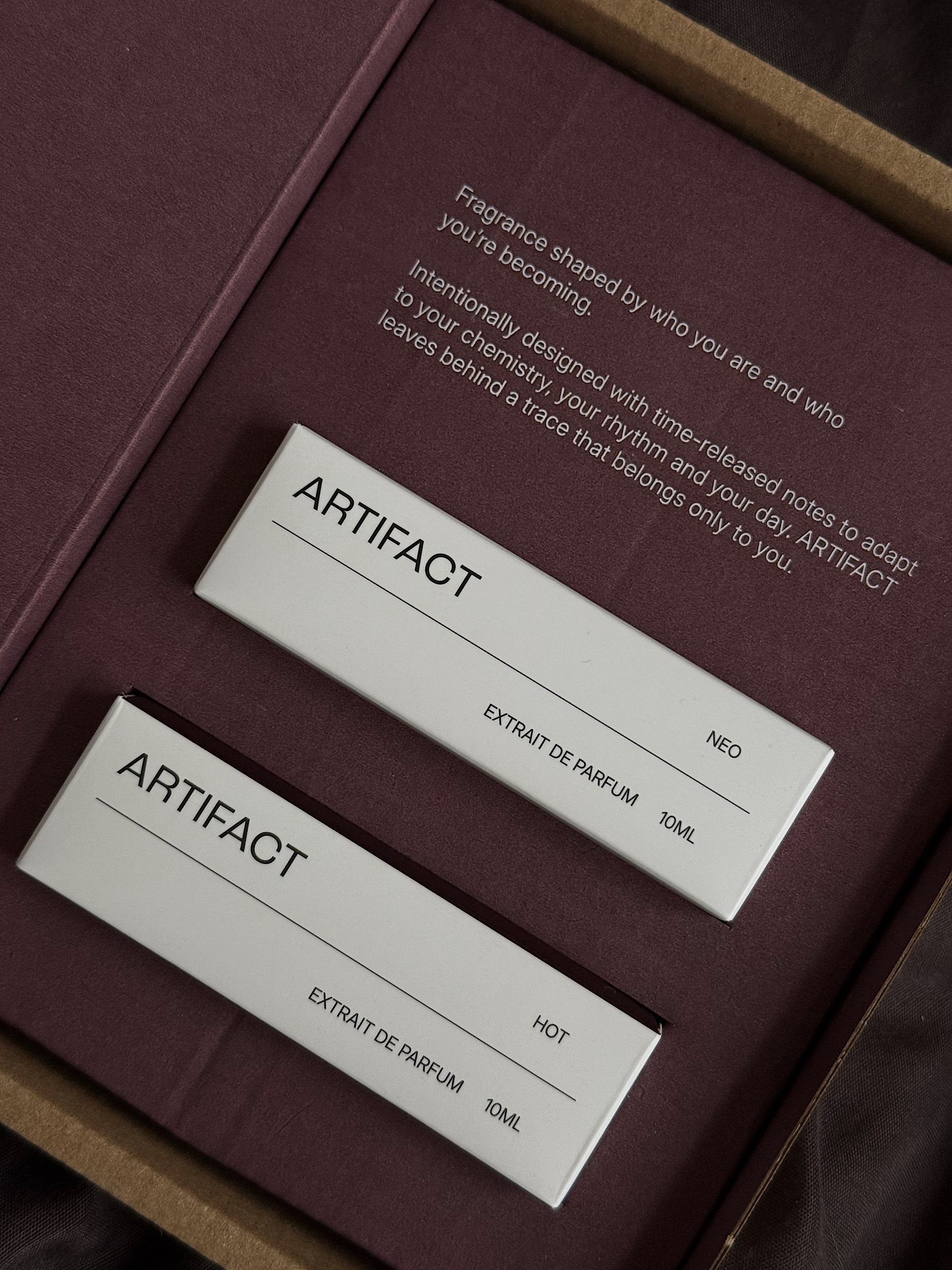 Two 'Artifact' perfume labels on a maroon box with text.